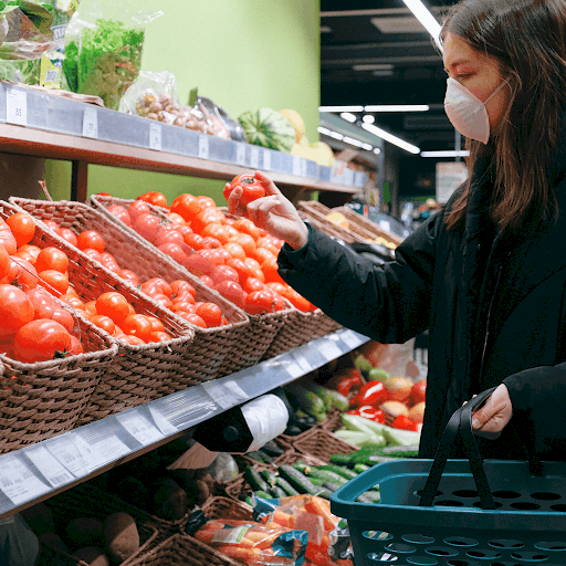 Woman wearing a mask holding a tomato shopping with grocery cart in hand