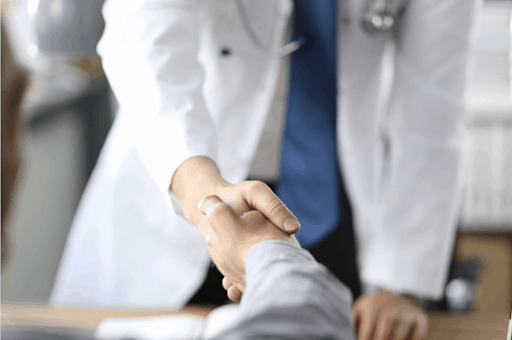 Doctor in white lab coat and stethoscope shaking hands with man sitting down at desk.