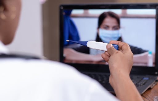  Female doctor in mask on computer screen reading temperature of patient with thermometer in hand at desk.