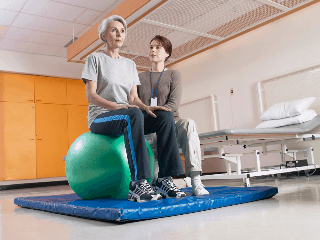 Woman on green ball next to physical therapist on blue mat